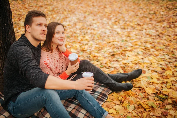 Hermosa pareja sentados juntos y mirar en una dirección. Tienen tazas de café. Él la abrazó. Se sientan en el parque . — Foto de Stock