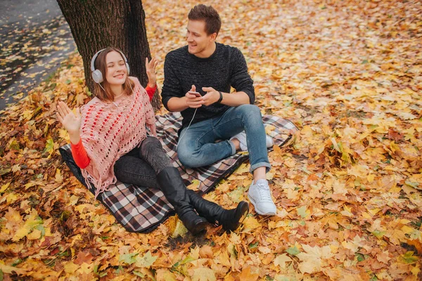 Jovem jovem e positiva gosta de ouvir música através de fones de ouvido. Ela senta-se no cobertor. O tipo está além dela. Ele segura o telefone e olha para a mulher . — Fotografia de Stock