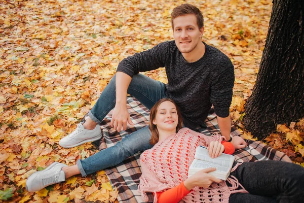 Hermosa pareja está en manta en el parque de otoño. Mujer joven acostada en piernas de chicos. Se sienta sobre una manta. Miran a Cemera y sonríen . — Foto de Stock