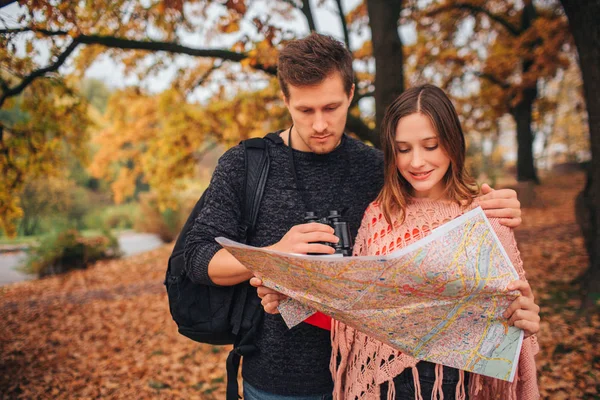 Concentrated young tourists stand in park and look at map. She holds it and smiles. People are in park. — Stock Photo, Image