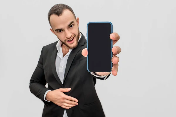 Confident young and bearded man in suit holds phone close to camera. He looks straight. Guy holds another hand on stomach. Isolated on white background. — Stock Photo, Image