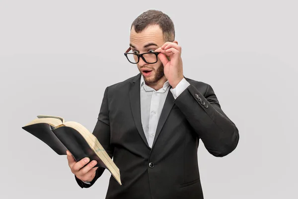 Je me demandais et excité jeune homme tenir livre dans une main et des lunettes avec un autre. Il regarde dans le livre. Il porte un costume. Isolé sur fond blanc . — Photo