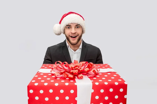 Amazed and excited young man hold big red present box. He looks on camera and smile. Guy wear Christmas hat and suit. Isolated on white background. — Stock Photo, Image