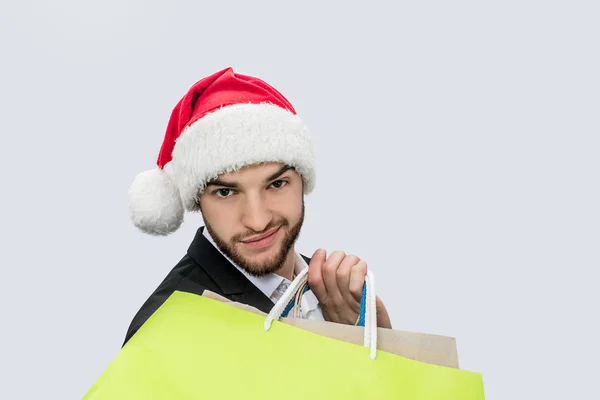 Young man in Christmas hat and suit hold yellow bag on shiulder and look on camera. He is confident. Isolated on white background. — Stock Photo, Image