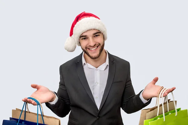 Un jeune homme joyeux et positif sourit à la caméra. Il porte un chapeau de Noël et un costume noir. Le jeune homme a des sacs à provisions dans les deux mains. Isolé sur fond blanc . — Photo