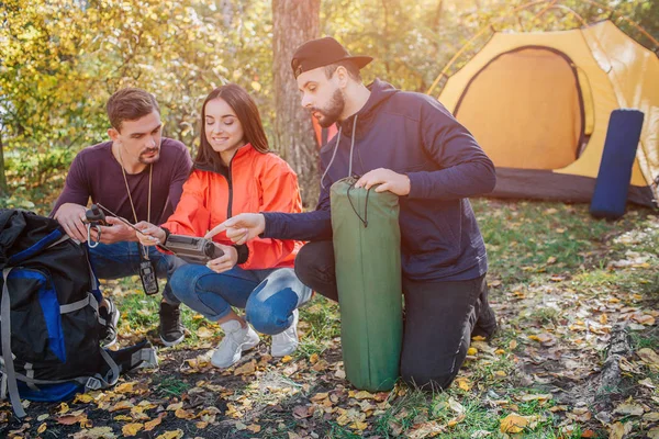 Three friends sit in squad position and look at radio young woman holds. Bearded guy holds sleeping bag. Another young man has knife in cover. They talk.
