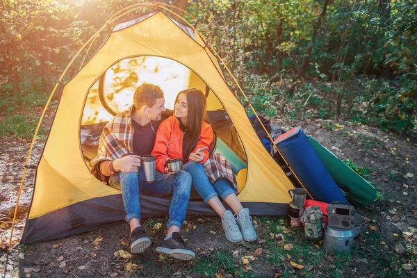 Позитивная молодая пара сидит в палатке и слушает музыку через одну пару наушников. Они смотрят друг на друга и улыбаются. Она держит телефон. У него термос. . — стоковое фото
