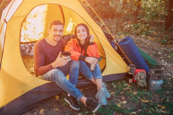 Preciosa imagen de pareja sentados juntos en la tienda y mirar en la cámara. Sonríen. La gente tiene termocopas. Hay mochila con equipo . — Foto de Stock