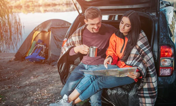 Tow oturist sit in trunk and look at map she holds. Young woman points on it. Guy smiles and holds cup. They are at lake.