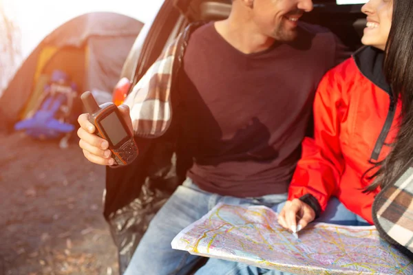 Cut view of lovely couple sitting together in trunk and smile. He holds satellite phone. She has map. People are close to tent. — Stock Photo, Image