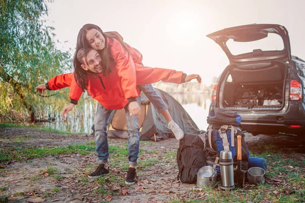 Cheerful young woman playing with guy. She sits on his back and look to right. Guy keeps hands aisde. They are at car, tent and lake. — Stock Photo, Image