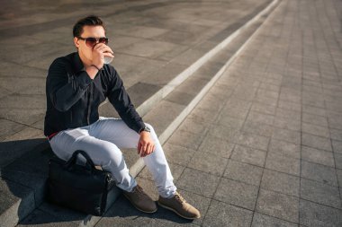 Sakin ve huzurlu genç adam gözlüklü merdivenlerinde dışarıda oturmak ve bekliyoruz. Bardaktan içer. Dışarıda hava güneşli.