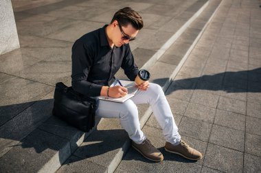 Konsantre genç adam adımları Caddesi ve yazma not defterinde oturamaz. O kahve elinde tutar. Adam neye gözlük sayesinde. Dışarıda hava güneşli.