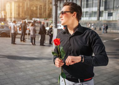 Siyah tişörtlü genç adam dışarıda dur, basılı tutarak ellerinde kırmızı gül. Bir kenara güneş gözlüğü ile görünüyor. Adam bekliyor. Onun arkasında insanlar var. Dışarıda hava güneşli.