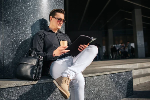 El hombre serio y concentrado se sienta afuera en los escalones y lee el diario. Tiene una taza de café en las manos. El tipo mira a través de gafas. Sostiene una pierna sobre otra. . — Foto de Stock