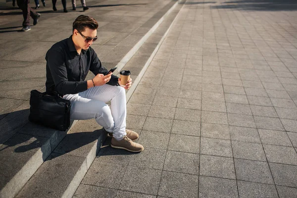 Goed geklede jonge man op stappen buiten zitten en kijken naar telefoon. Hij houdt de kop koffie. Leerzak is naast hem. Is het zonnig is buiten. — Stockfoto