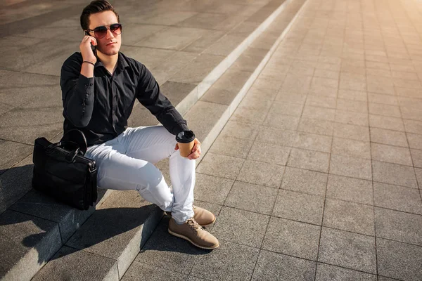 Beau jeune homme dans des lunettes parler au téléphone. Il regarde à travers des lunettes. Il tient une tasse de café à la main. Il est assis dehors sur les marches . — Photo