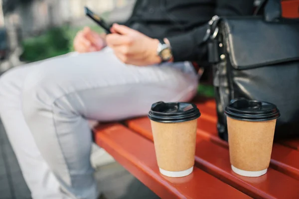 Cut vuew of man sitting on bench. There are cup nd black leather bag. He holds phone in hands. Guy sit outside.