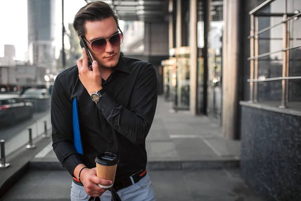 Jeune homme en lunettes de soleil marche à côté de la construction et parler au téléphone. Il tient la tasse et le sac dans une main. Il est occupé. . — Photo