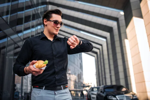 Un jeune homme occupé se tient debout et regarde les montres à travers des lunettes de soleil. Il tient en main un croissan salé. Il sourit un peu. . — Photo