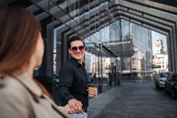 Un jeune homme joyeux se tient dehors avec une femme et lui tient la main. Il la regarde et sourit. Il tient une tasse de boisson et sourit. Elle le regarde. . — Photo