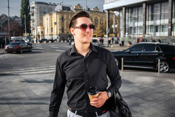 Beau et heureux jeune homme marche dans la rue. Il regarde à travers les lunettes de soleil. Le jeune homme a une tasse de boisson et un sac en cuir. Il est confiant . — Photo