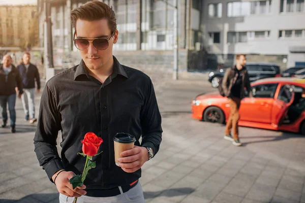 Un beau jeune homme se tient dehors. Il tient une rose rouge et une tasse de boisson. Il porte des lunettes de soleil. Il y a des gens et des voitures derrière lui . — Photo