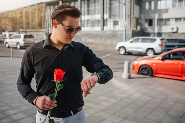 Le jeune homme se tient dehors dans la rue et tient la rose rouge. Il regarde les montres. Le type attend. Il y a une route avec des voitures derrière lui . — Photo