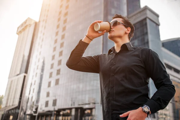 Photo de jeune homme debout devant le gratte-ciel et boire dans une tasse en papier. Il pose sur la caméra . — Photo