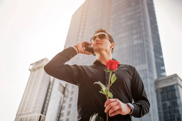 Un homme d'affaires se tient devant un gratte-ciel et parle au téléphone. Il tient la rose rouge en main. Il fait beau dehors . — Photo