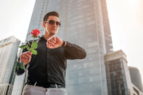 Jeune homme beau debout devant le gratte-ciel et regardez les montres. Il tient rose rouge dans la main . — Photo