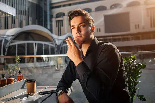 Photo d'un jeune homme assis à table et fumant. Il lève les yeux vers la droite. Il s'appuie sur la table. Il est assis dehors. Le soleil brille . — Photo