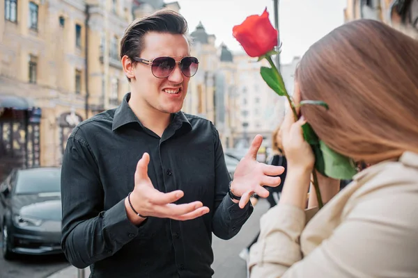 Young man stand outside in front of woman and talk to her. She holds red rose in hands. Guy wear sunglasses. — Stock Photo, Image