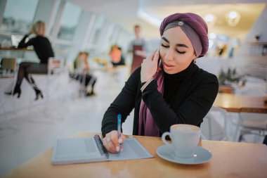 Genç Arap işkadını içinde masaya oturup not defterinde yazmak. Aynı anda telefonla konuşuyor. Modeli sit masada yalnız.