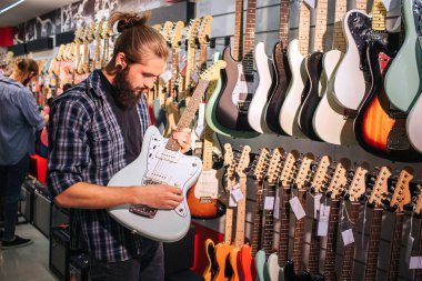 Sakallı genç adam stand ve gitar çalmak. Onu elinde tutmak. Orada başka bir elektrik gitar duvarda asılı.