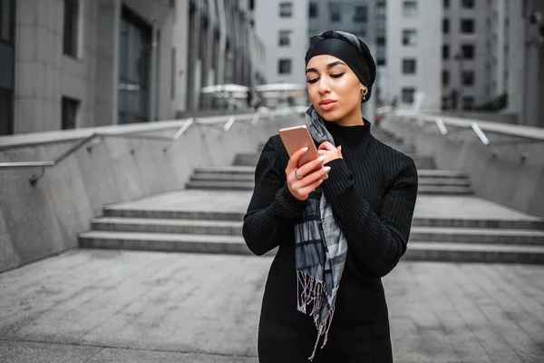 Beautiful young arabian woman stands and look on phone. She poses. Model wears hijab.