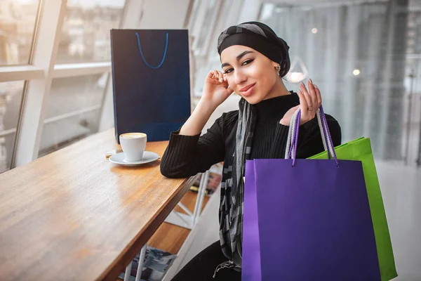 Bonita joven modelo árabe posando en cámara. Se sienta a la mesa cerca de la ventana del café. Joven mujer mantenga bolsas de compras con la mano . — Foto de Stock