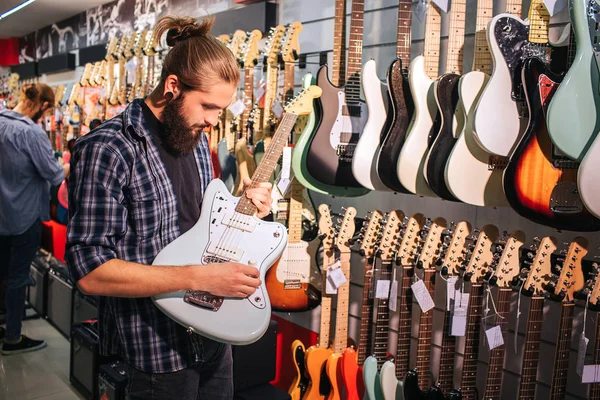 Young bearded man stand and play guitar. He hold it in hands. There are another electrical guitars hanging on wall.