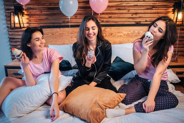 Tre unga glada kvinnor sitta på sängen. Två av dem titta på KANTPRES och leende. Tredje äter man den. Kaka är läckra. Flickor är i rummet. Vissa baloons är bakom. — Stockfoto