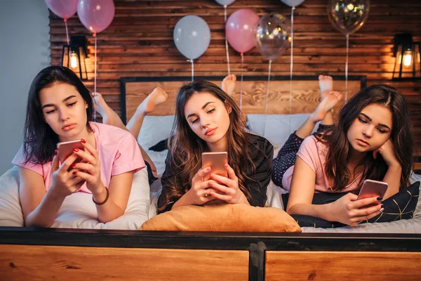 Ernsthafte und geschäftige Austernfrauen, die auf dem Bett im Zimmer liegen. Sie halten Telefone in der Hand und schauen sie an. Mädchen sind traurig und konzentriert. — Stockfoto