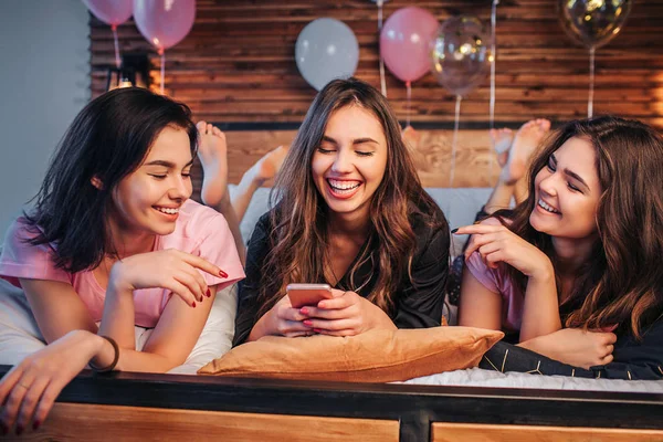 Fröhliche und glückliche junge Frauen, die im festlichen Raum auf dem Bett liegen. sie lächeln. Mädchen in der Mitte halten Telefon in den Händen. zwei weitere Modelle zeigen auf Gerät. — Stockfoto