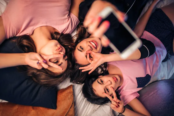 Três jovens divertidas deitadas na cama. Eles posam na câmera e fazem poses diferentes. Modelo no meio segurar telefone branco na mão . — Fotografia de Stock