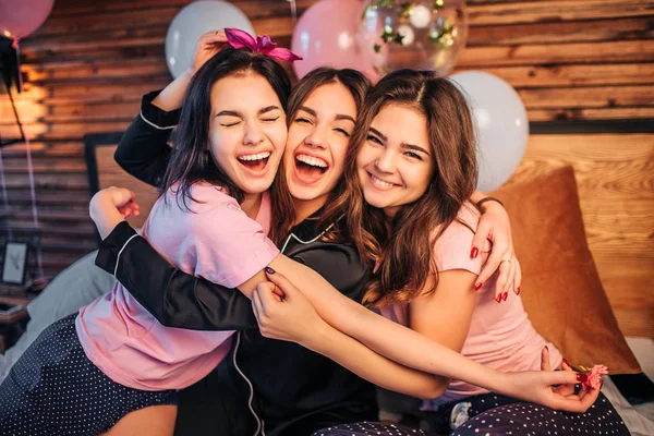 Jovens modelos alegres e felizes abraçando uns aos outros. Eles se sentam na cama no quarto juntos e olham para a câmera. Os adolescentes usam pijama. Eles têm festa em casa . — Fotografia de Stock