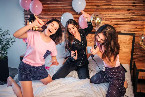 Jovens divertidas e simpáticas, ajoelhadas na cama do quarto. Fingem cantar em microfones. Modelos segurar escova de maquiagem, lata de pulverização e equalizador de cabelo nas mãos . — Fotografia de Stock