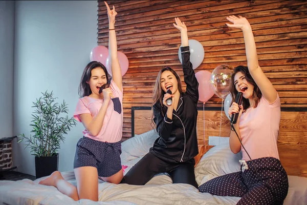 Drei junge Models haben Spaß auf dem Bett im Zimmer. Sie geben vor, in Dingen zu singen, die sie in Händen halten. Mädchen halten die Hände hoch. sie schreien und singen. — Stockfoto
