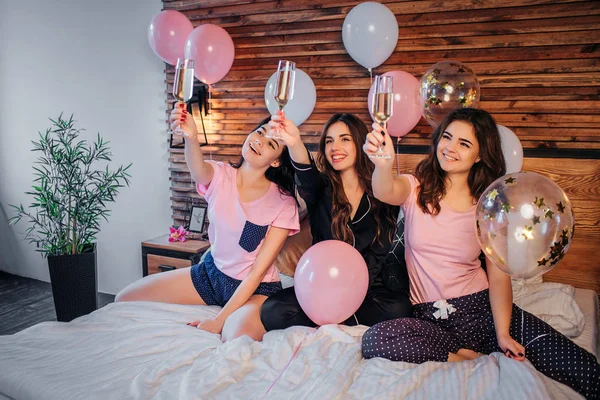 Alegre jóvenes modelos se sientan en la cama en la habitación festiva. Sujetan las copas con champaña en el aire y las miran. Chica sonriendo. Tienen fiesta de pijamas. . —  Fotos de Stock