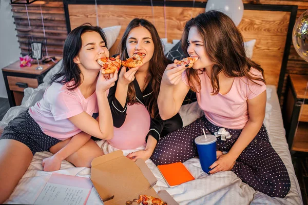 Drie hongerig tieners bijten stukken uit pizza. Ze verslinden het. Hongerige modellen dragen pyjama's. Ze zitten op bed in kamer. — Stockfoto