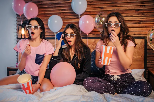 Förvånade och glada unga kvinnor sititng på sängen i festlig rum och titta flyttar. De wea speciella glasögon. Flickor är glada. Två modeller håller hinkar med popcorn. Tredje har man cup med cola. — Stockfoto