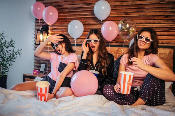 Unga kvinnor har discgussed känslor på ansiktet. De titta på film via speciella glasögon. Modeller sitter på sängen i festlig rum. De har pyjamas part. — Stockfoto