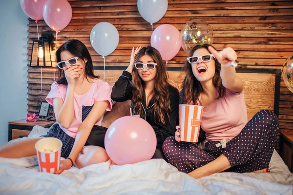 Emotionale junge Frauen sitzen auf dem Bett in einem festlichen Raum. Sie sehen Filme durch spezielle Brillen. Models tragen Pyjamas. Sie haben Party gemacht. — Stockfoto
